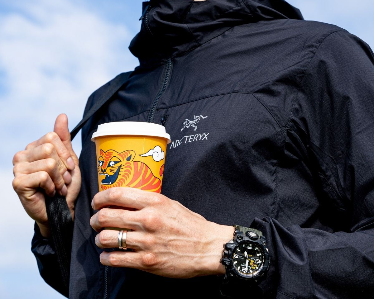 A person holding a specialty coffee cup while wearing an Arc'teryx jacket, representing the union of specialty coffee and nature.